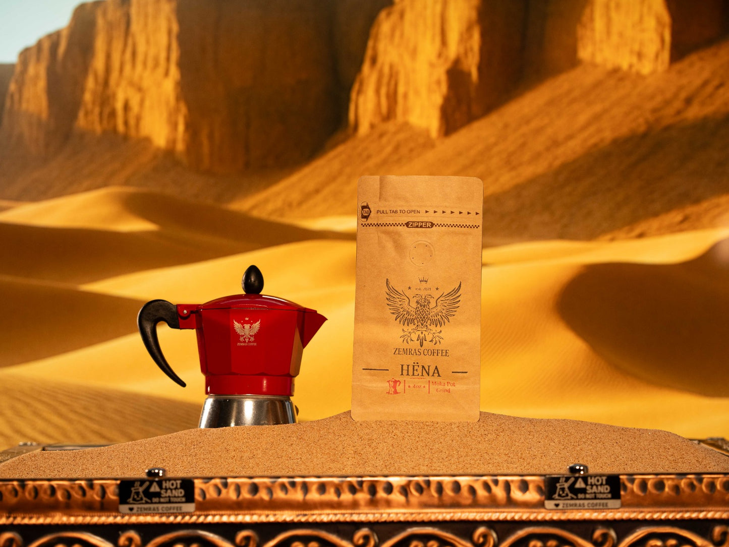 Hëna "Moon" decaf coffee bag and a red Moka pot on sand, featuring a fine grind for a rich stovetop espresso ritual.