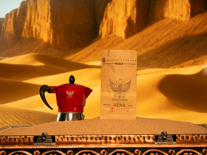 Hëna "Moon" decaf coffee bag and a red Moka pot on sand, featuring a fine grind for a rich stovetop espresso ritual.