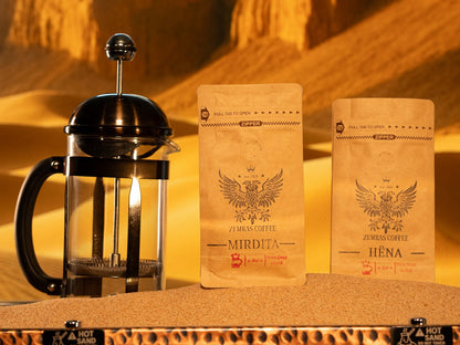 Mirdita and Hëna coffee bags with a French press on sand, featuring specific coarse grinds for caffeinated and decaf rituals.