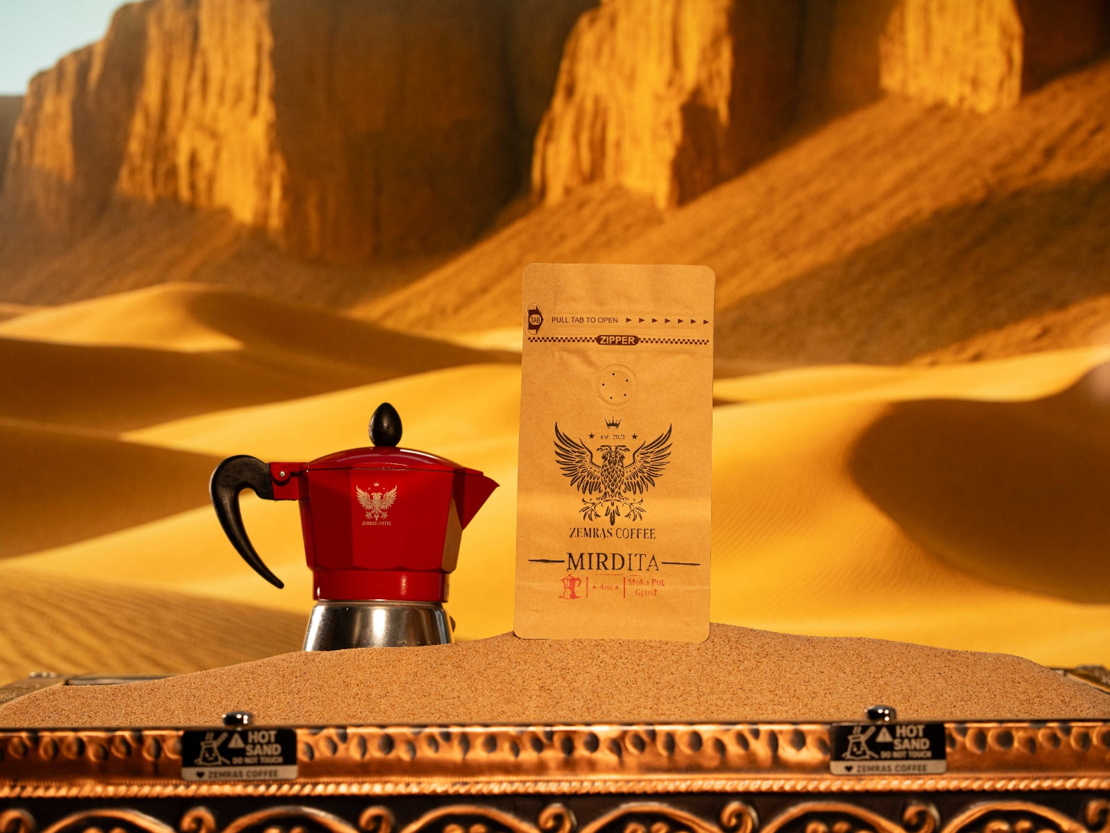 Mirdita "Good Day" Colombian dark roast coffee bag and a red Moka pot on a sand-brewing station for authentic stovetop espresso.