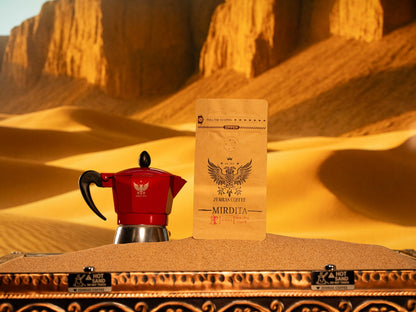Mirdita "Good Day" Colombian dark roast coffee bag and a red Moka pot on a sand-brewing station for authentic stovetop espresso.